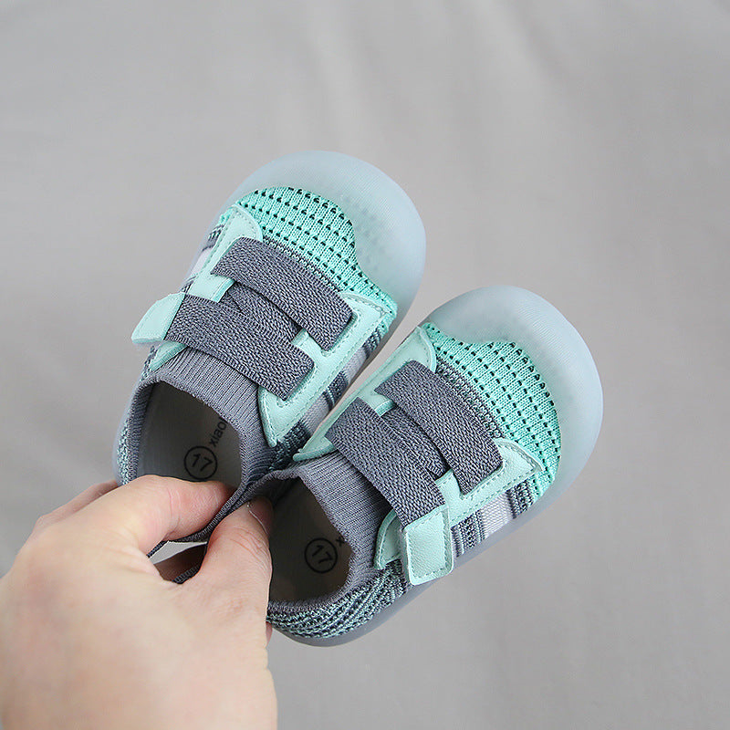 A hand holds a pair of small children's shoes. The shoes are light blue and gray with mesh fabric and elastic straps, set against a neutral background.