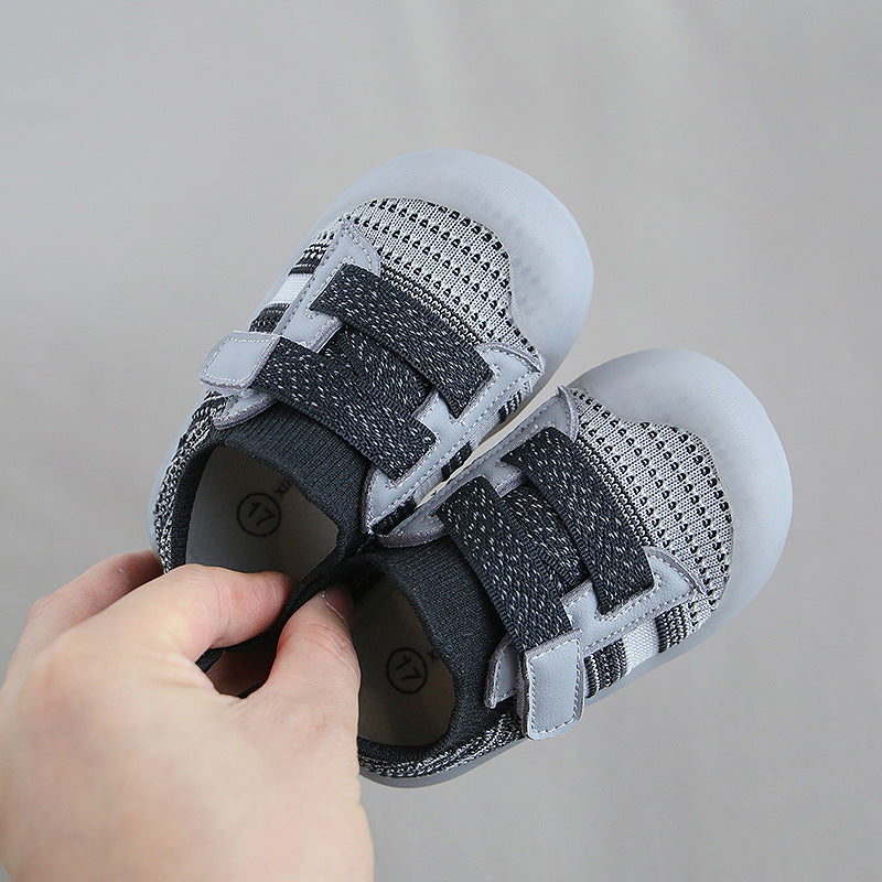 A hand holding a pair of small, gray baby shoes with black and white mesh design, featuring two velcro straps and light soles, against a plain gray background.