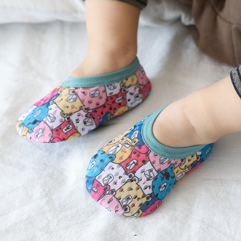 Baby wearing colorful slippers with cartoon animal faces, including bears and cats. Slippers feature a teal trim and are worn on a white fabric surface.