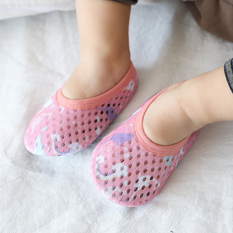 Baby feet wearing pink, perforated slip-on shoes with a playful pattern. The shoes rest on a light fabric, highlighting the comfort and breathable design.