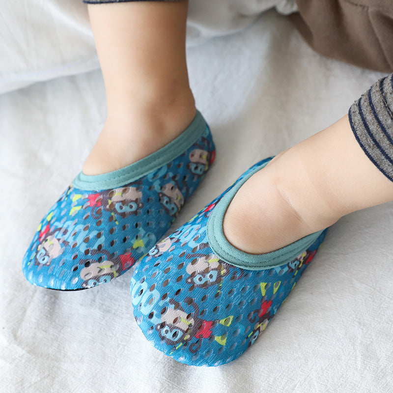 Close-up of a child's feet wearing blue, breathable slippers with playful cartoon faces and colorful patterns. The slippers have teal accents and are on a white fabric surface.