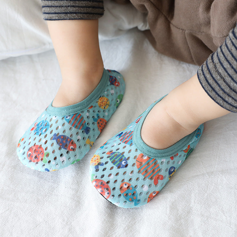 Toddler wearing blue mesh baby shoes with colorful fish patterns, sitting on a white sheet. The child also wears striped grey leggings.