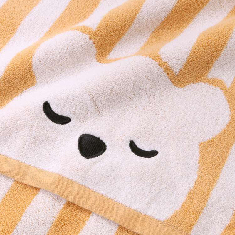 Close-up of a towel featuring a sleeping bear face with closed eyes. The towel has a soft texture and alternating beige and yellow stripes in the background.