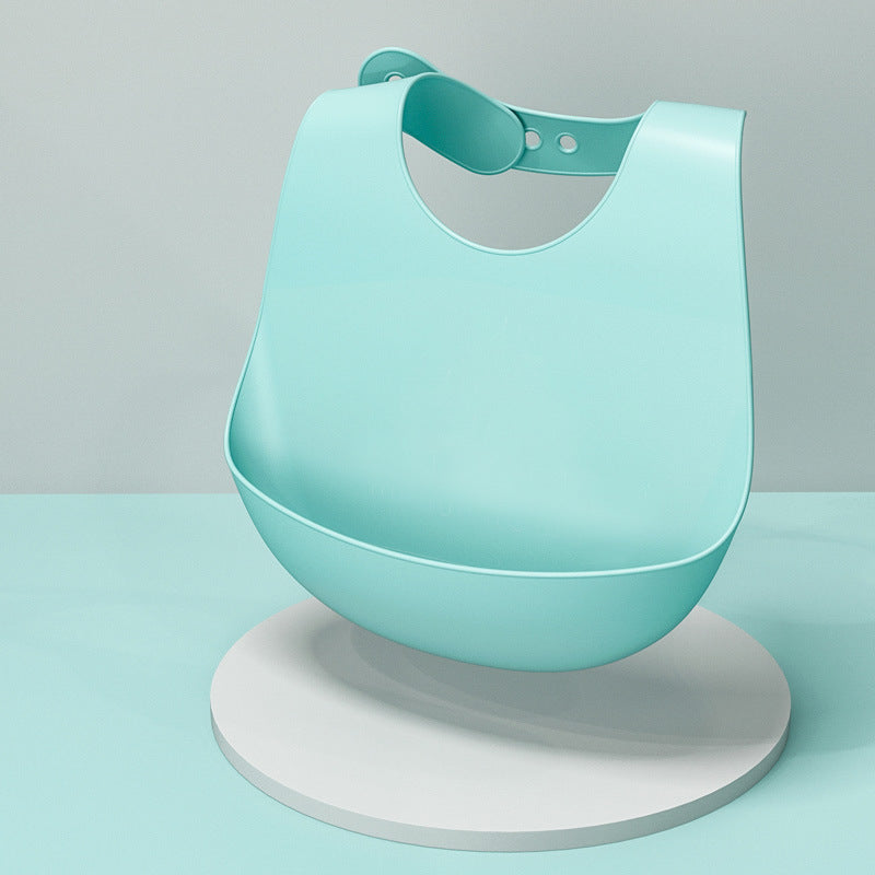 A light blue, silicone baby bib with an adjustable neck strap and a wide food-catching pocket is displayed on a round white podium against a pastel background.