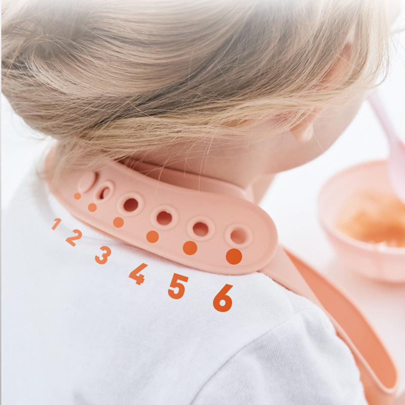 A child wearing an adjustable pink silicone bib with numbered holes on the strap for sizing. The child is facing away, next to a bowl of orange-colored food on a white surface.