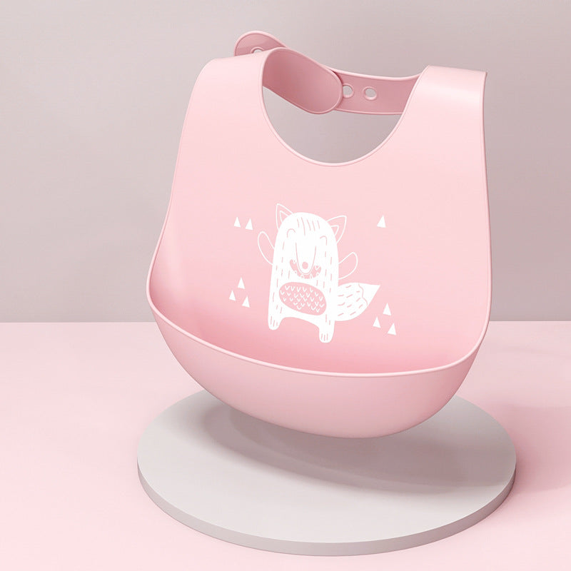 A pink silicone bib with an illustration of a smiling fox in the center, surrounded by small triangles. The bib is on a round white stand against a soft pink background.
