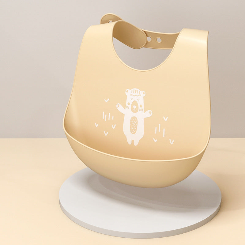 A soft beige silicone baby bib with a white bear illustration in the center. The bib has an adjustable strap and is placed upright on a round, light-colored base.