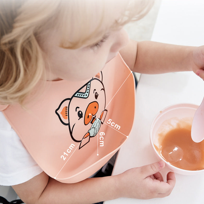 Toddler wearing a peach-colored bib with a pig design is eating from a bowl of orange puree with a spoon. Measurements of the bib are shown as 21cm, 5cm, and 6cm.