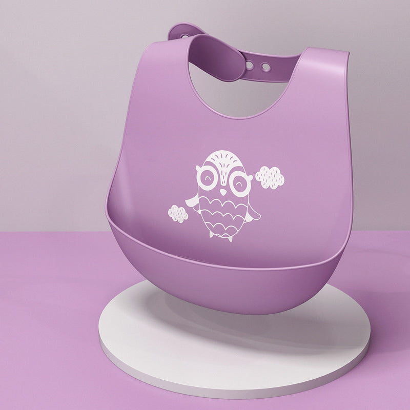 Purple baby bib with an owl and clouds design, displayed on a circular white stand against a pink background. Fastened with adjustable buttons at the neck.