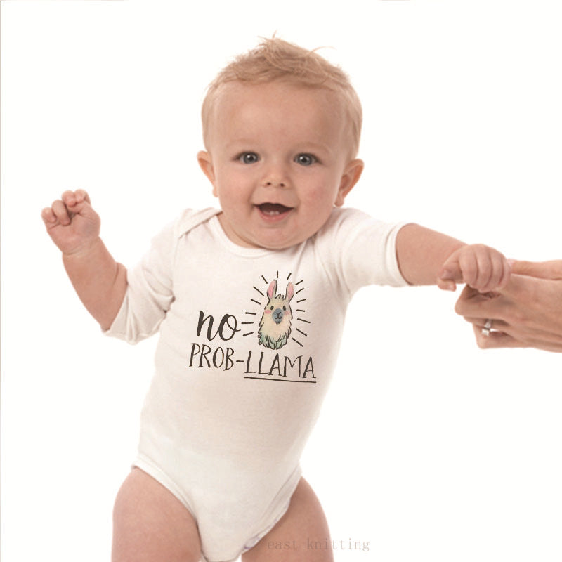 A smiling baby wearing a white onesie with the text "no prob-llama" and a drawing of a llama. An adult's hands support the baby who appears to be learning to stand.