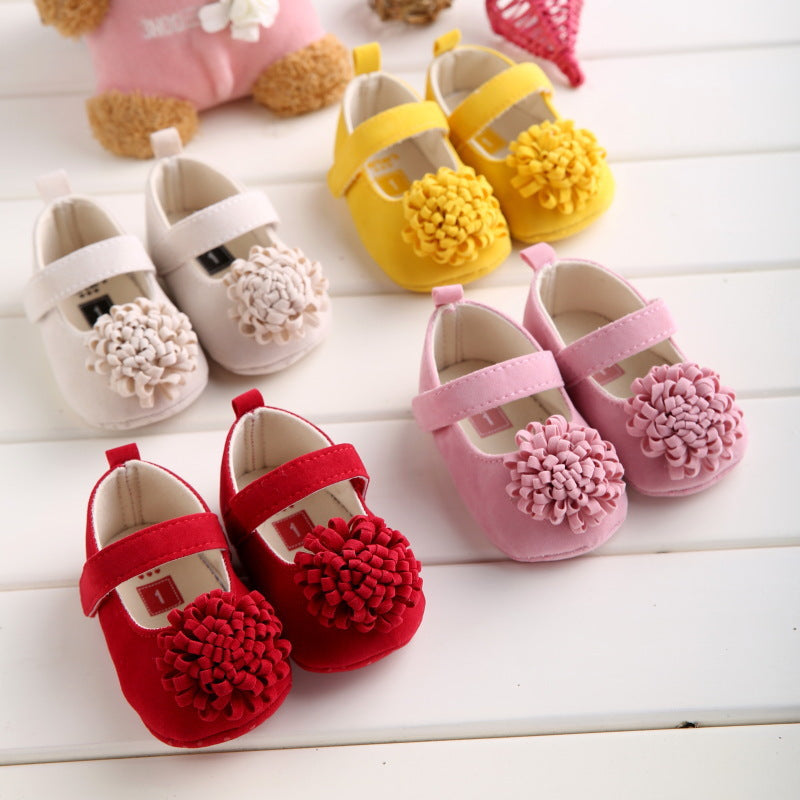 Four pairs of baby shoes with floral decorations are arranged on a white surface. The shoes are white, yellow, pink, and red, each featuring a large flower embellishment.