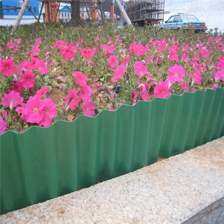 Pink flowers bloom behind a green corrugated metal fence in an urban setting, with a stone curb in the foreground. A parked car and buildings are visible in the background.