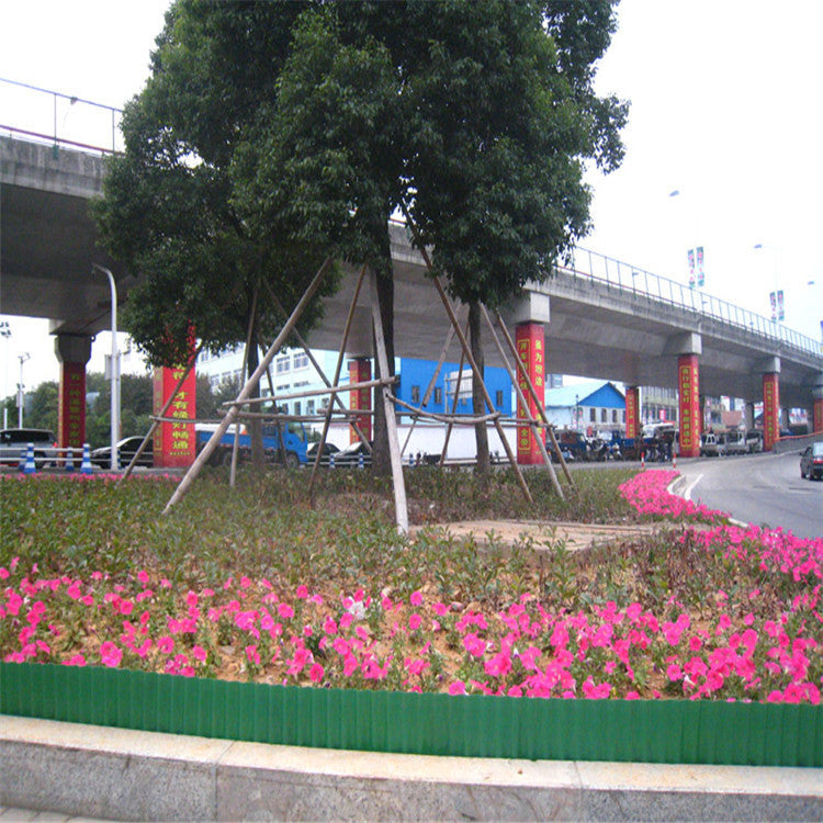 Pink flowers and lush greenery border a curved road under a large overpass. Vehicles are seen in the background beneath the elevated highway.