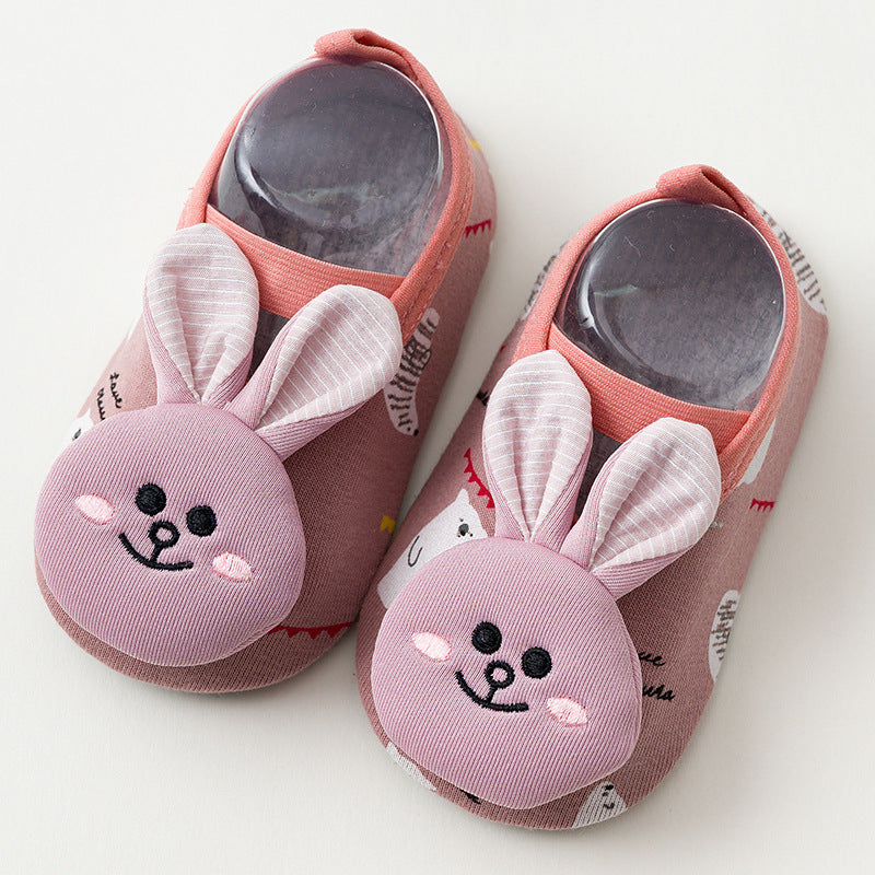 Pink baby slippers with cute bunny faces featuring embroidered eyes and smiles, and soft fabric ears. The slippers have a patterned design and are shown from above.