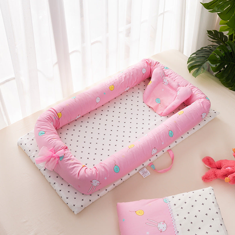 Pink baby nest with star-patterned cushion and pillow, featuring colorful doodles. A pink ribbon is tied to the side. Sunlight filters through sheer curtains.