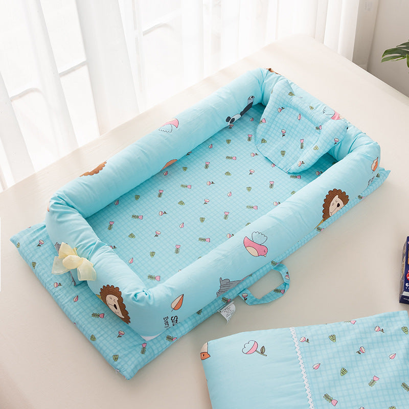 Light blue baby nest with cute animal and flower patterns, featuring soft padded edges and a small pillow. It rests on a white surface near sheer curtains.