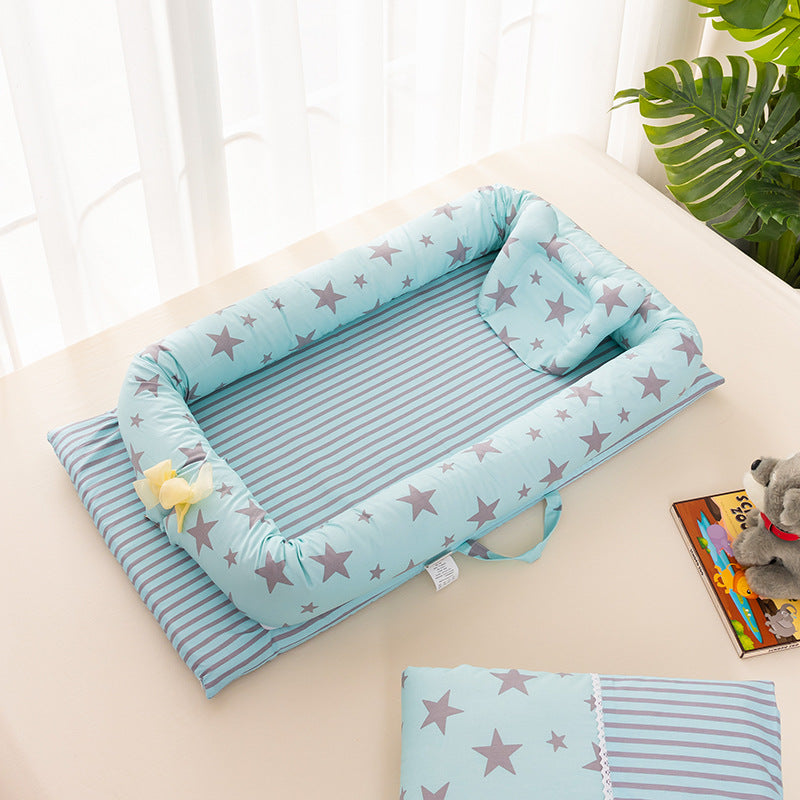 Light blue baby nest with star and stripe patterns, placed on a table beside a toy, book, and cushion. Nearby are sheer curtains and a green plant for a cozy setup.