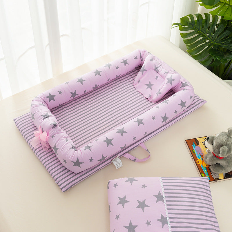 A pink baby nest with gray stars and striped pattern on a bed, accompanied by a matching pillow and a plush toy. A large green plant adds a touch of nature in the background.