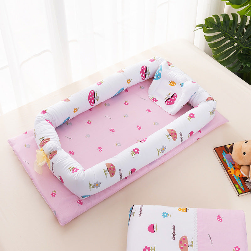 A pink baby play mat with a cushioned border featuring colorful mushroom and flower prints, placed on a light surface. A teddy bear and a book are nearby.