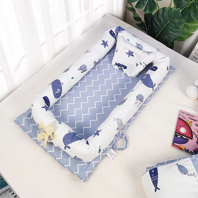 Blue and white baby nest with ocean-themed prints on a chevron-patterned mattress. It rests on a bed next to a plant and a toy book.