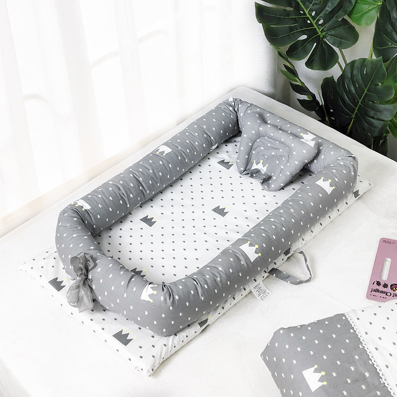 Gray baby nest with crown patterns on a polka-dotted mattress, placed on a white surface. A leafy green plant in the corner adds a touch of nature.