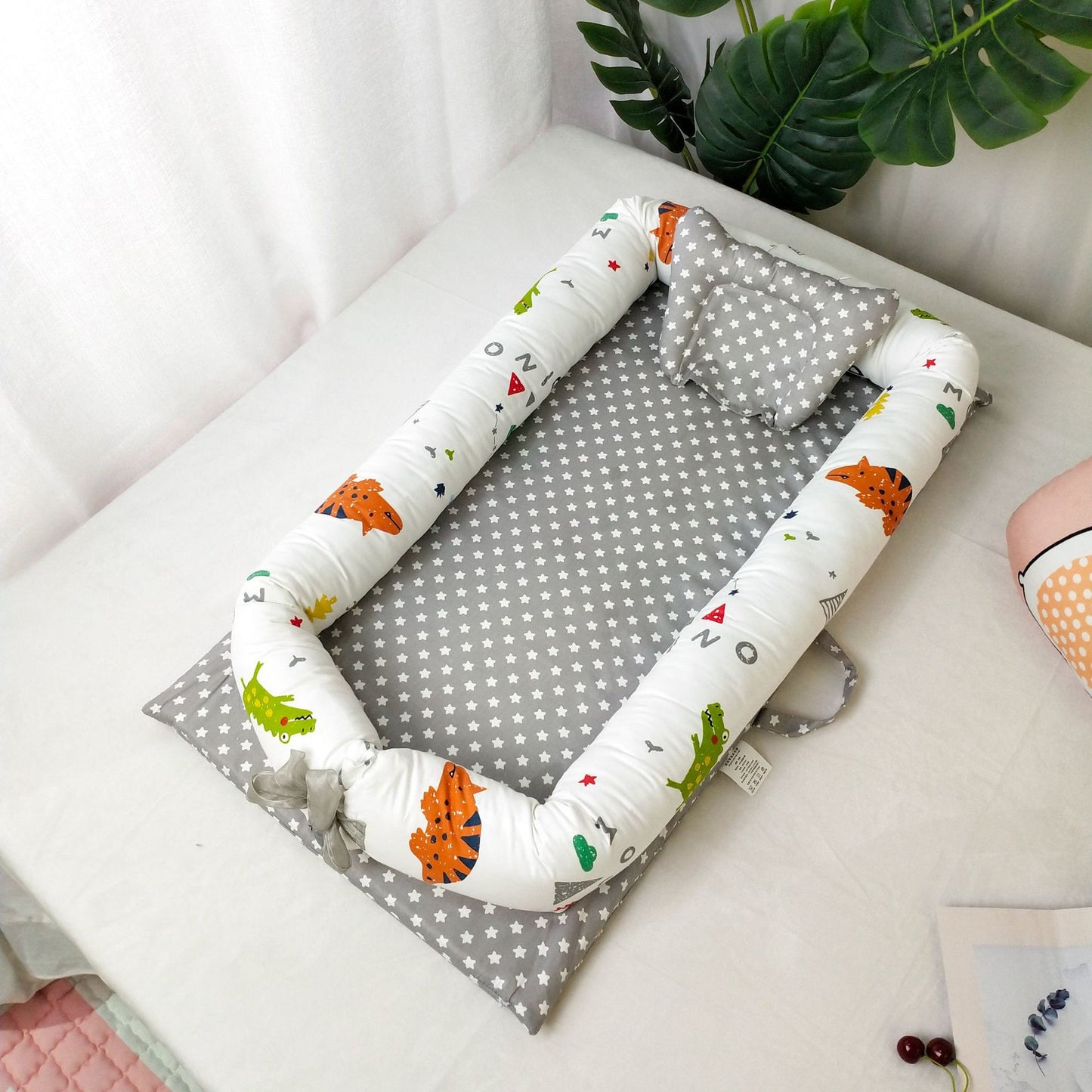 Baby nest with a gray star-patterned base and colorful dinosaur designs on the padded sides. There's a matching pillow, all placed on a light-colored surface.