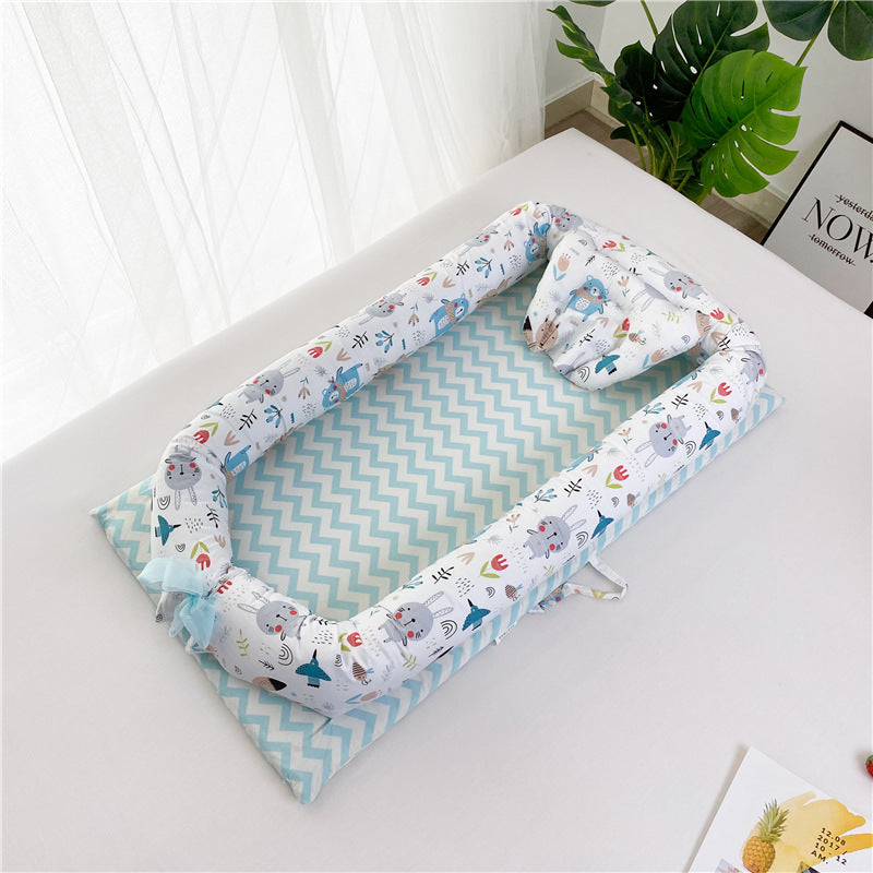 Baby nest with chevron-patterned mattress and printed animal designs on the sides and pillow. It's placed on a white surface near a large green plant and a decorative frame.