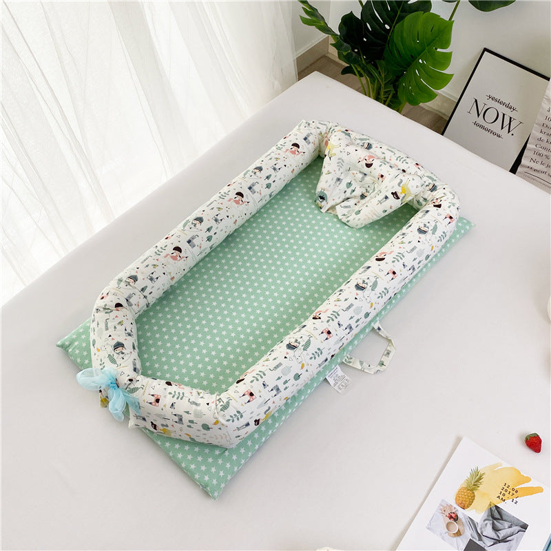 Infant bed with a mint green, star-patterned mattress and cartoon-print cushioned bumpers on a light surface near a leafy plant and framed prints.