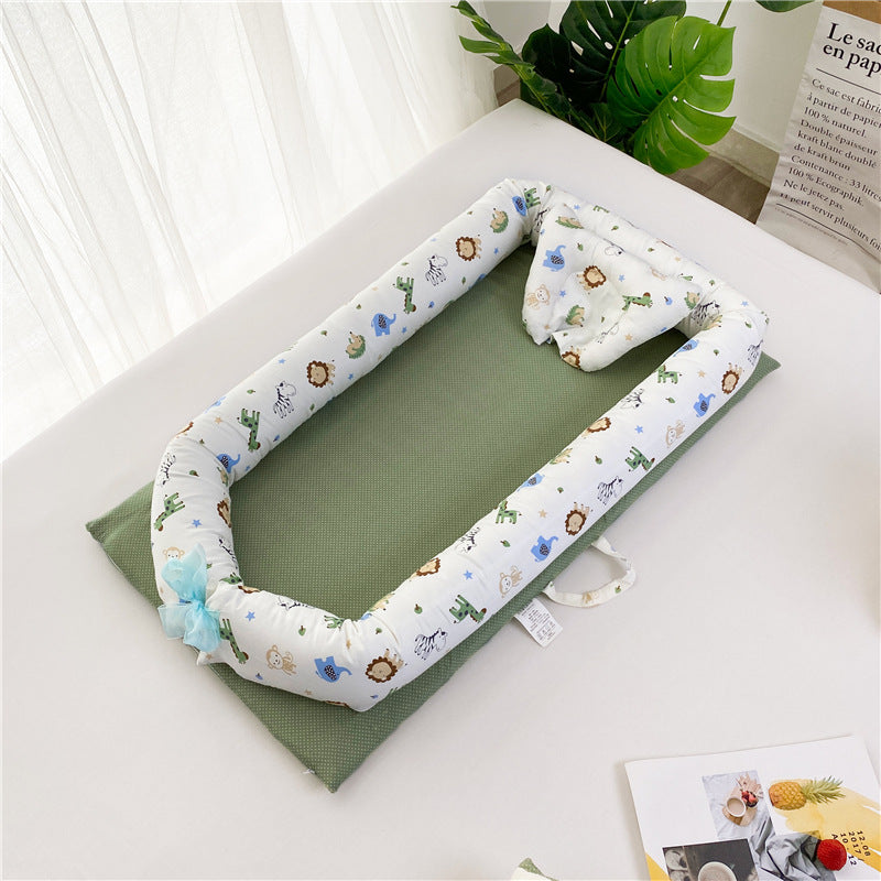 Baby nest with a green base and a white padded border featuring animal patterns. It's placed on a white bed next to a large leaf, a booklet, and sheer curtains.