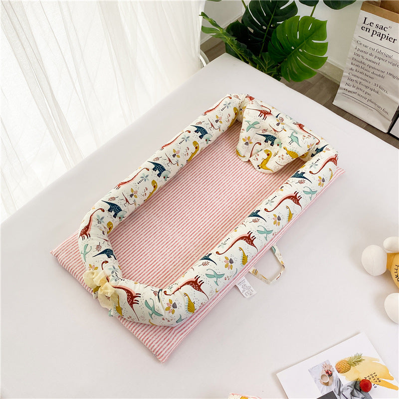 Baby lounger with a pink striped base and dinosaur-patterned cushioned sides. It rests on a white surface, with decorative plants and a paper bag in the background.