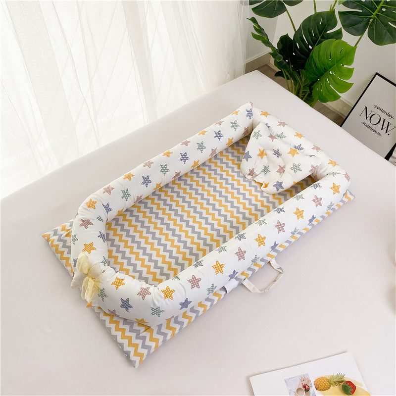 Baby nest with a colorful star pattern, chevron inner fabric, and matching pillow on a light surface next to a potted plant and pineapple-themed magazine.