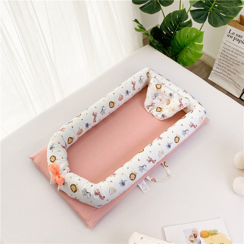 A pink baby lounger with a white, animal-patterned border, featuring lions and elephants. It sits on a table near green plants and softly lit by natural light.