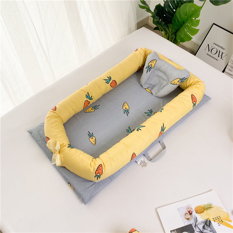 Yellow and gray baby cocoon with carrot patterns, set on a white surface near a large green plant and a wall featuring framed art. Soft light filters through a nearby window.