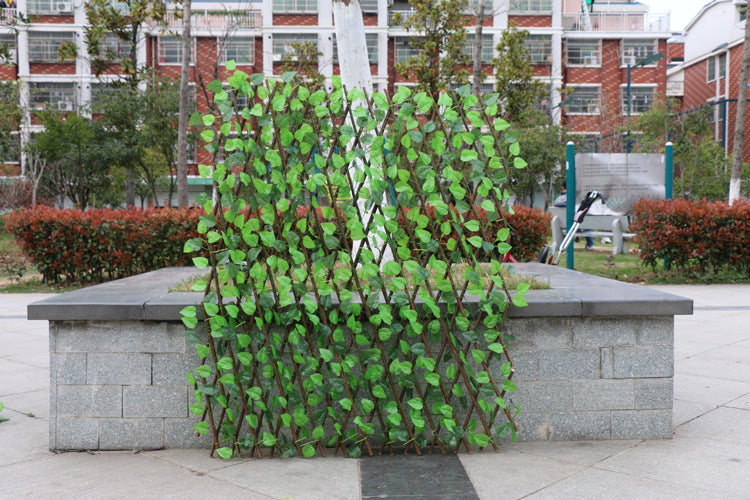 A lattice screen with green leaves is placed in front of a stone bench in a park. The background features a red brick building and surrounding greenery.