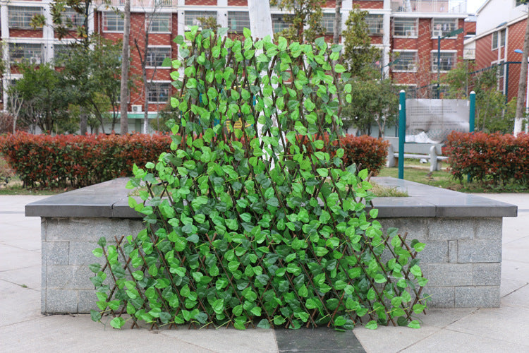 A decorative trellis with vibrant green leaves leans against a stone bench in a courtyard. Red brick buildings and bushes in the background complete the urban setting.