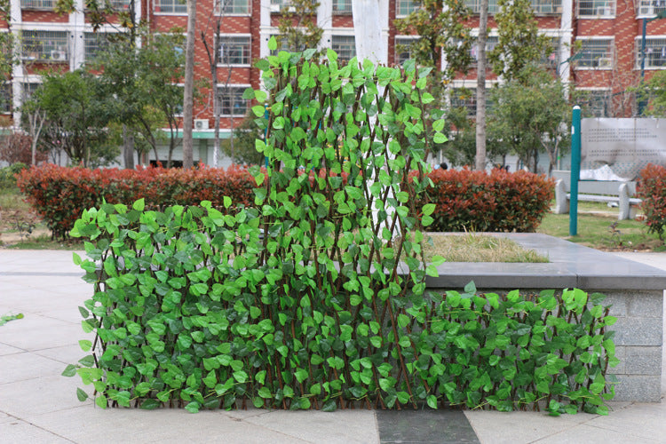 A park setting shows a bench covered with a lattice of green leaves, blending with the surrounding red bushes and trees. A brick building is visible in the background.