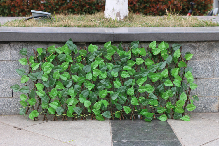 Expandable green leaf trellis against a gray stone wall with grass on top. The trellis is vibrant green, contrasting with the muted stone and concrete surfaces around it.