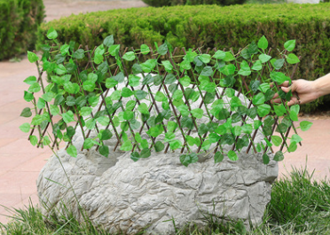 Expandable trellis with green leaves stretched over a large rock, surrounded by grass. A hand adjusts the trellis. Shrubs and a paved pathway are visible in the background.