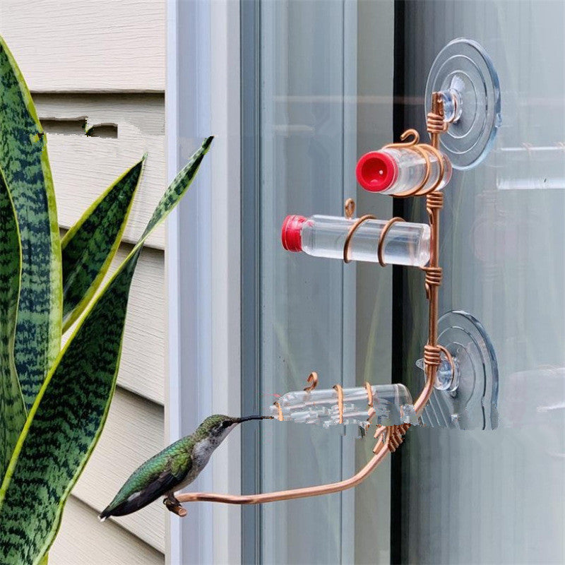 A hummingbird drinks from a feeder attached to a glass window. The feeder consists of tubes with red lids held by copper wire, near a plant with long, pointed leaves.