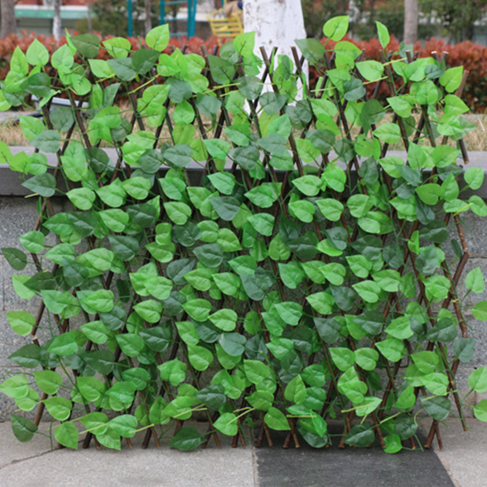 Artificial green leaf trellis with lush, vibrant foliage arranged on a wooden lattice. It's placed outdoor against a stone surface with blurred park background.