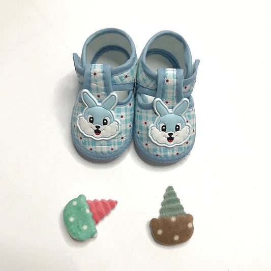 Blue baby shoes with smiling bunny faces on a plaid pattern, placed on a white background. Two toy ice cream cones, one green and pink, the other green and brown, are in front.