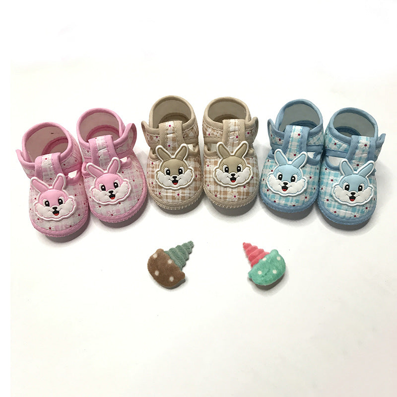 Three pairs of baby shoes—pink, beige, and blue—with cartoon bunny faces displayed on a white background. Two small plush ice cream cone toys are placed in front.