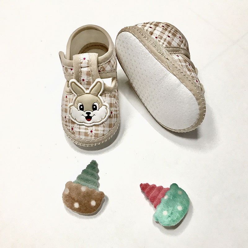 Plaid baby shoes with bunny design, featuring one shoe upside down to show its white sole. Two crocheted ice cream cone toys in brown and green, and pink and green lie nearby.
