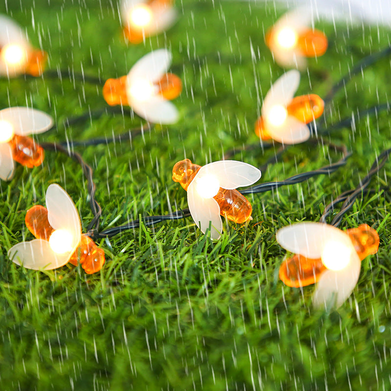 String lights shaped like bees with glowing bulbs adorn a grassy surface in the rain, creating a whimsical and enchanting outdoor atmosphere.