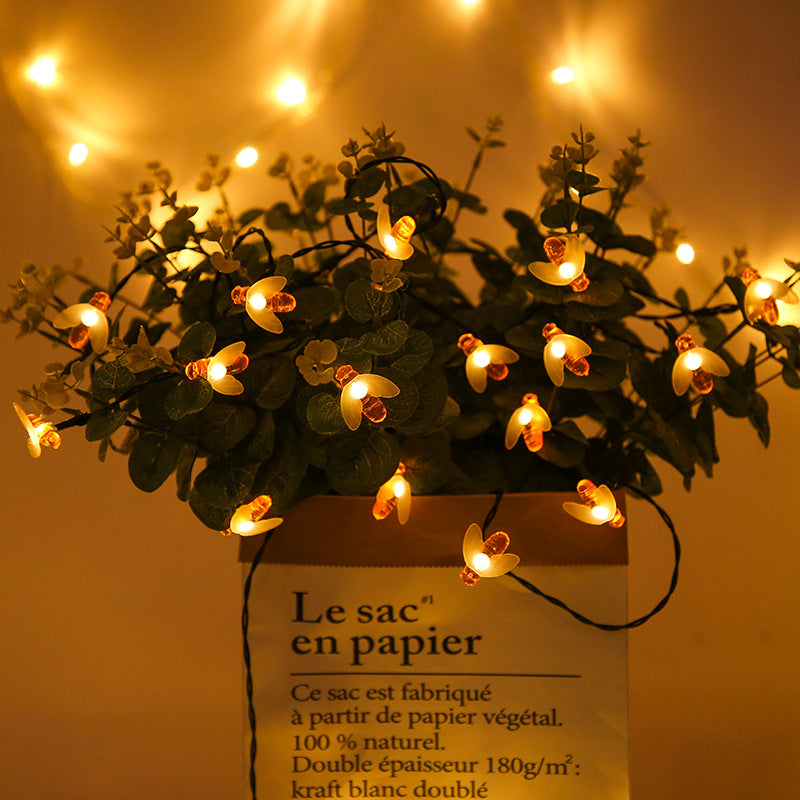 String lights shaped like bees illuminate a potted plant with lush green leaves in a paper bag, creating a warm, cozy ambiance with a glowing background.