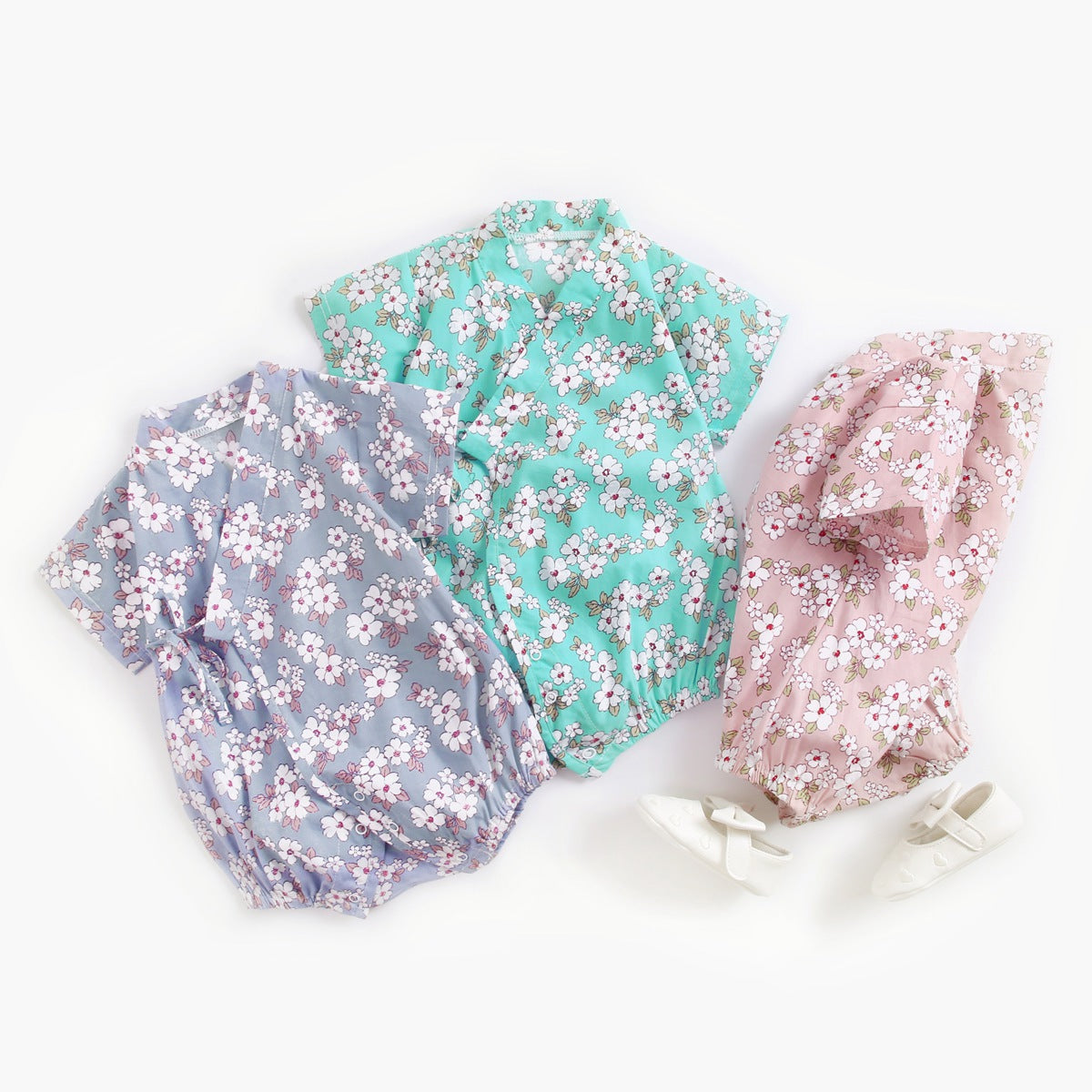 Three floral-patterned baby rompers in blue, mint, and pink are laid out on a white background, accompanied by a pair of white baby shoes.