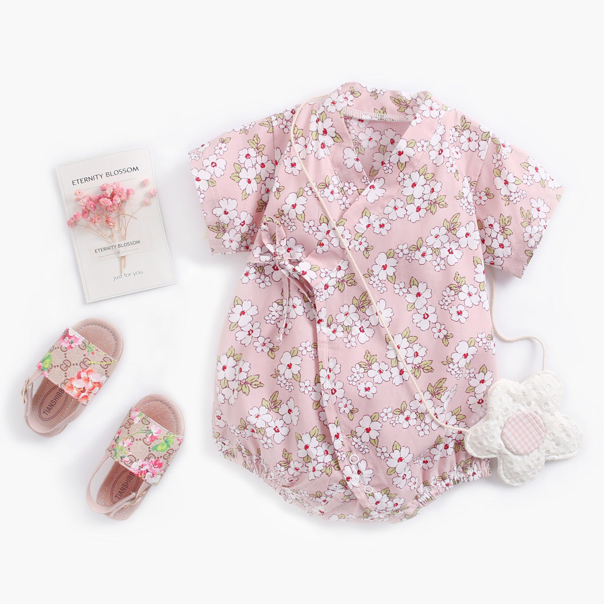 Pink floral baby romper with short sleeves, matching flower-shaped teether, floral sandals, and small card with dried pink flowers on a white background.