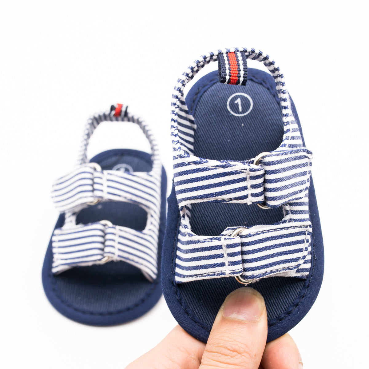 Navy blue baby sandals with white and blue striped straps, featuring buckle details, held up by a hand, against a plain white background.