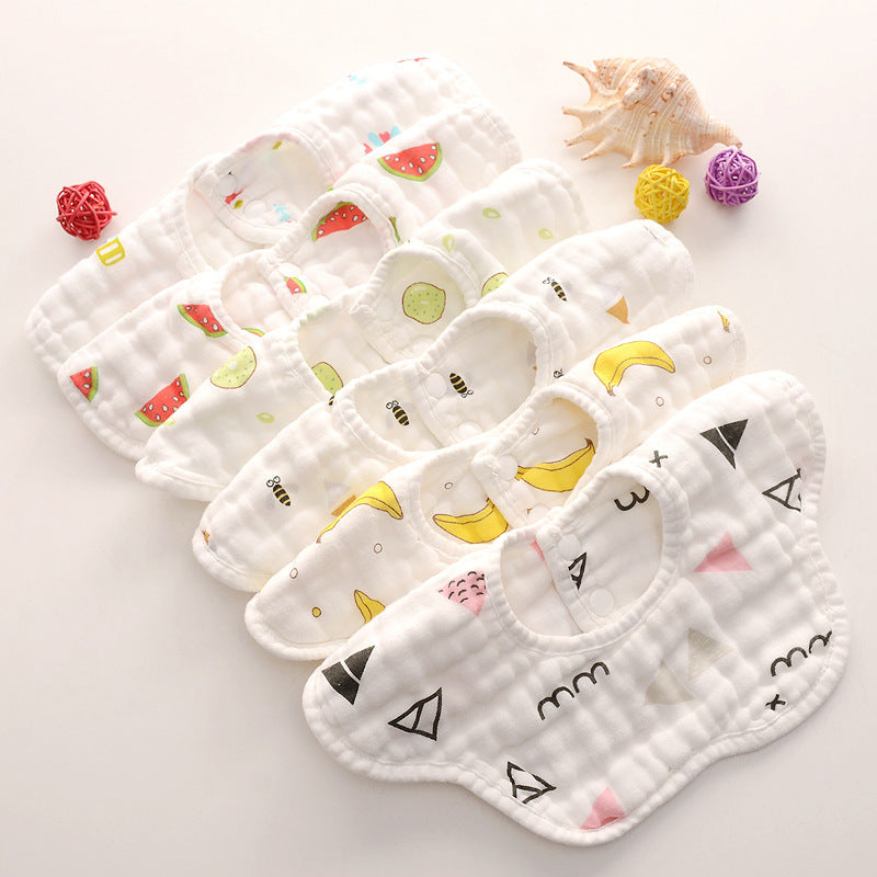 A collection of seven white baby bibs with various colorful prints, including watermelons, cucumbers, bees, bananas, and geometric shapes, arranged neatly on a light surface.
