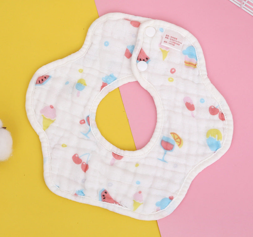A flower-shaped baby bib with colorful patterns of ice cream, fruits, and drinks lies on a pink and yellow background. It has snap buttons and a soft, quilted texture.